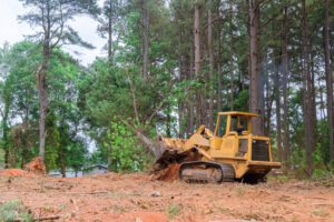Yellow bulldozer with a front bucket clearing soil on a dirt plot among tall trees in a forested area.