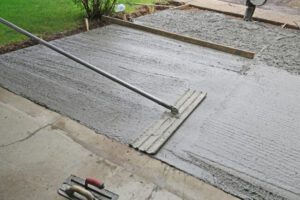 Fresh concrete is being spread and leveled with a long screed across a newly poured slab outside, with a trowel tool nearby.